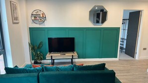 En stue med en grøn sofa, et fladskærms-TV på et træstel, en grøn accentvæg, en lille reol med dekorative genstande, og et spejl på væggen.