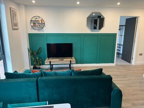 A living room with a green sofa, a flat-screen TV on a wooden stand, a green accent wall, a small shelf with decorative items, and a mirror on the wall. - Superb citycentre new built flat and gym (York)