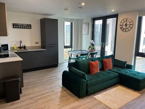 A modern kitchen with dark cabinets, a dining area with a table and chairs, and a green sectional sofa with orange pillows. - Superb citycentre new built flat and gym (York)