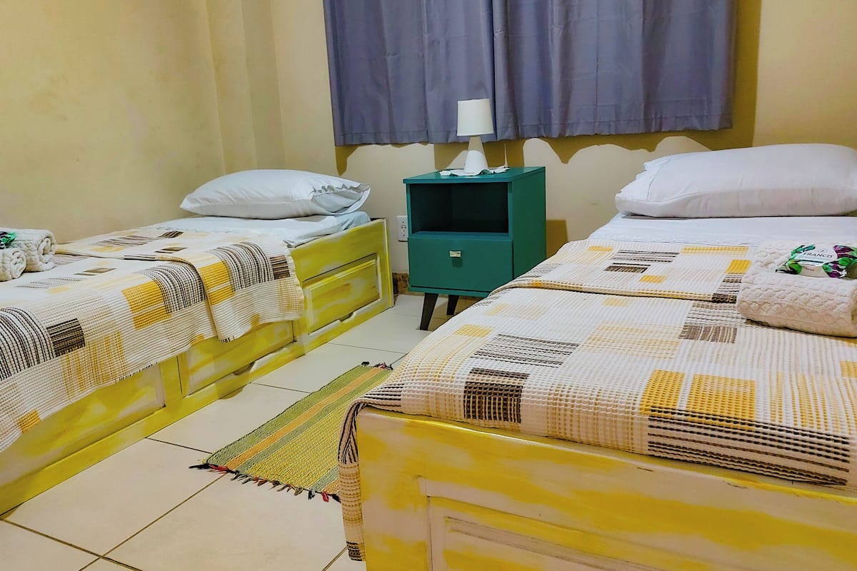 A hotel room with two beds, each with a white pillow and a rolled towel. There is a green nightstand with a lamp and a small table.