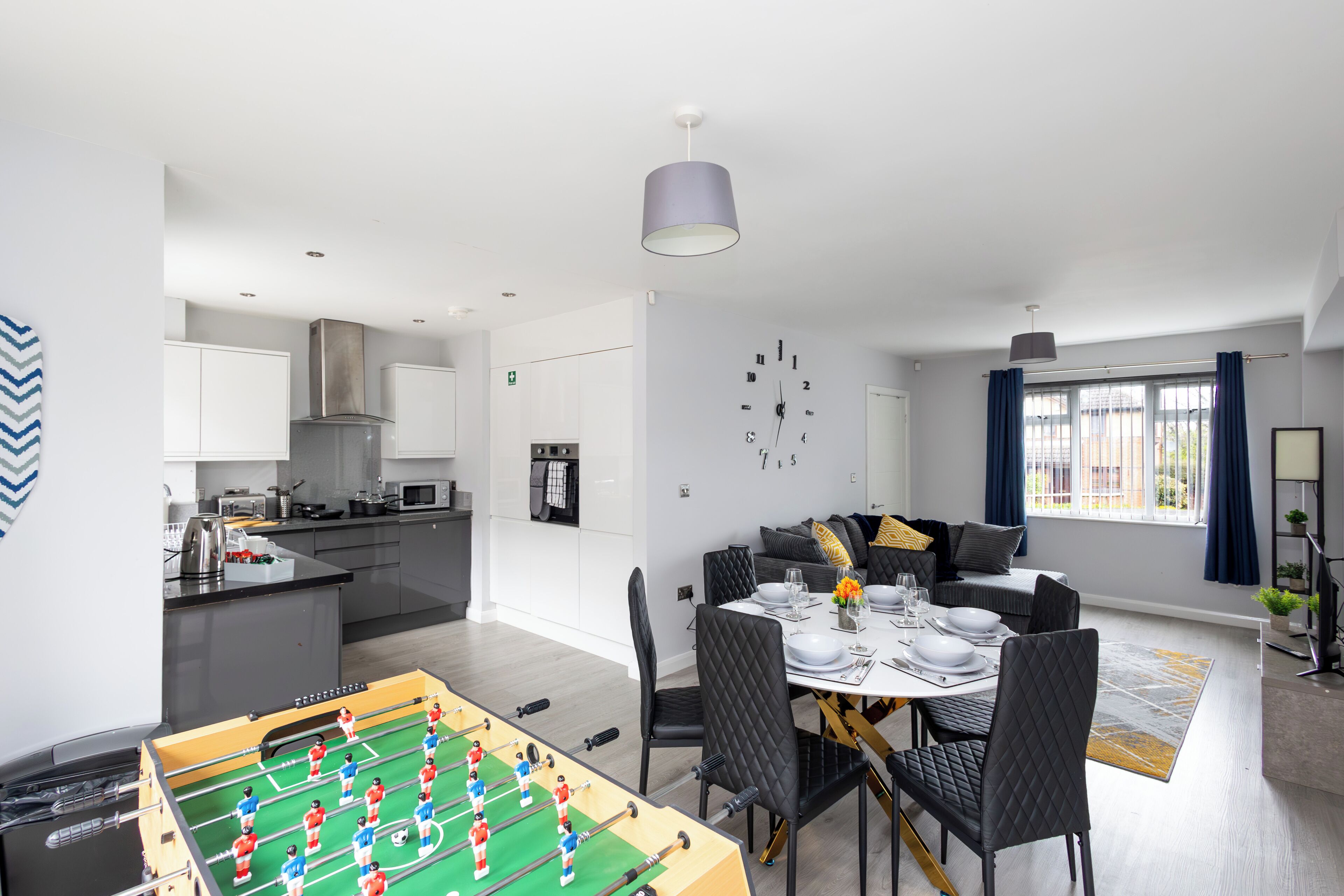 A modern kitchen with a foosball table, dining area with a table set for four, and a living area with a sofa and a clock on the wall.