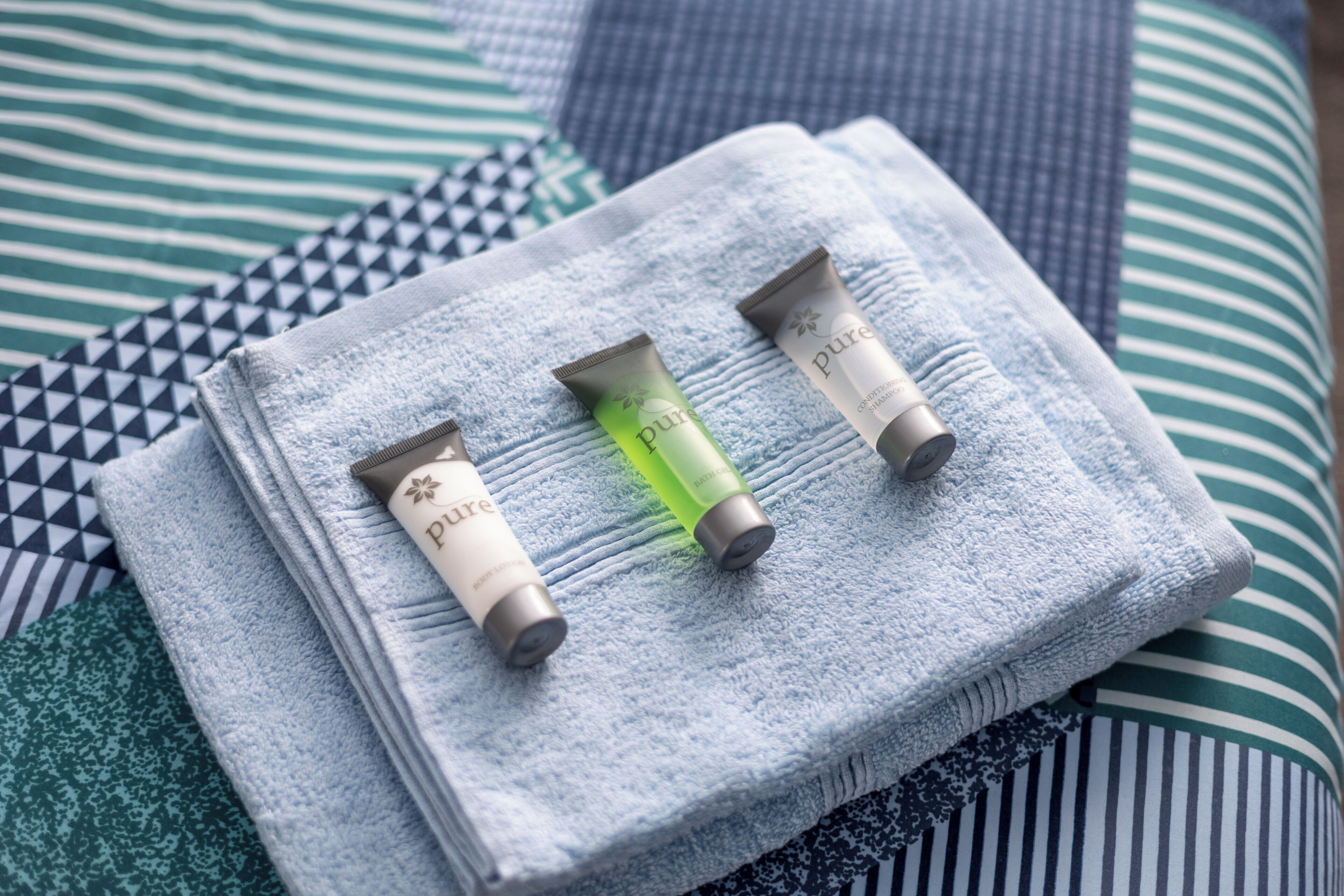 Une serviette soigneusement pliée, accompagnée de trois tubes de gel douche Pure, est posée sur un lit dont la literie est à rayures.