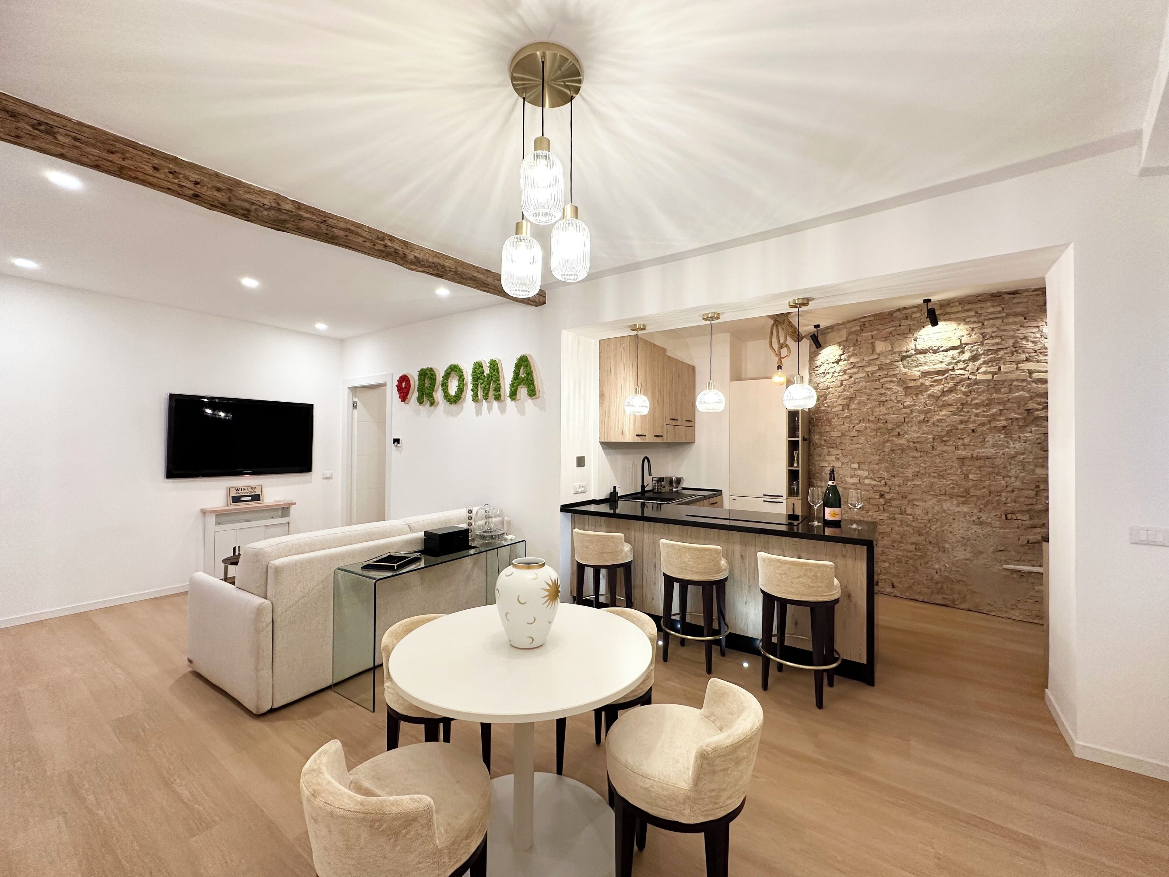 A modern kitchen with a stone wall backsplash, a dining area with a round table and chairs, and a living area with a sofa and a TV.