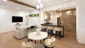 A modern kitchen with a stone wall backsplash, a dining area with a round table and chairs, and a living area with a sofa and a TV. - House in the shadow of the Colosseum (Roma)