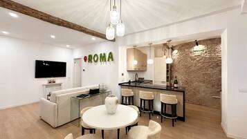 A modern kitchen with a stone wall backsplash, a dining area with a round table and chairs, and a living area with a sofa and a TV.