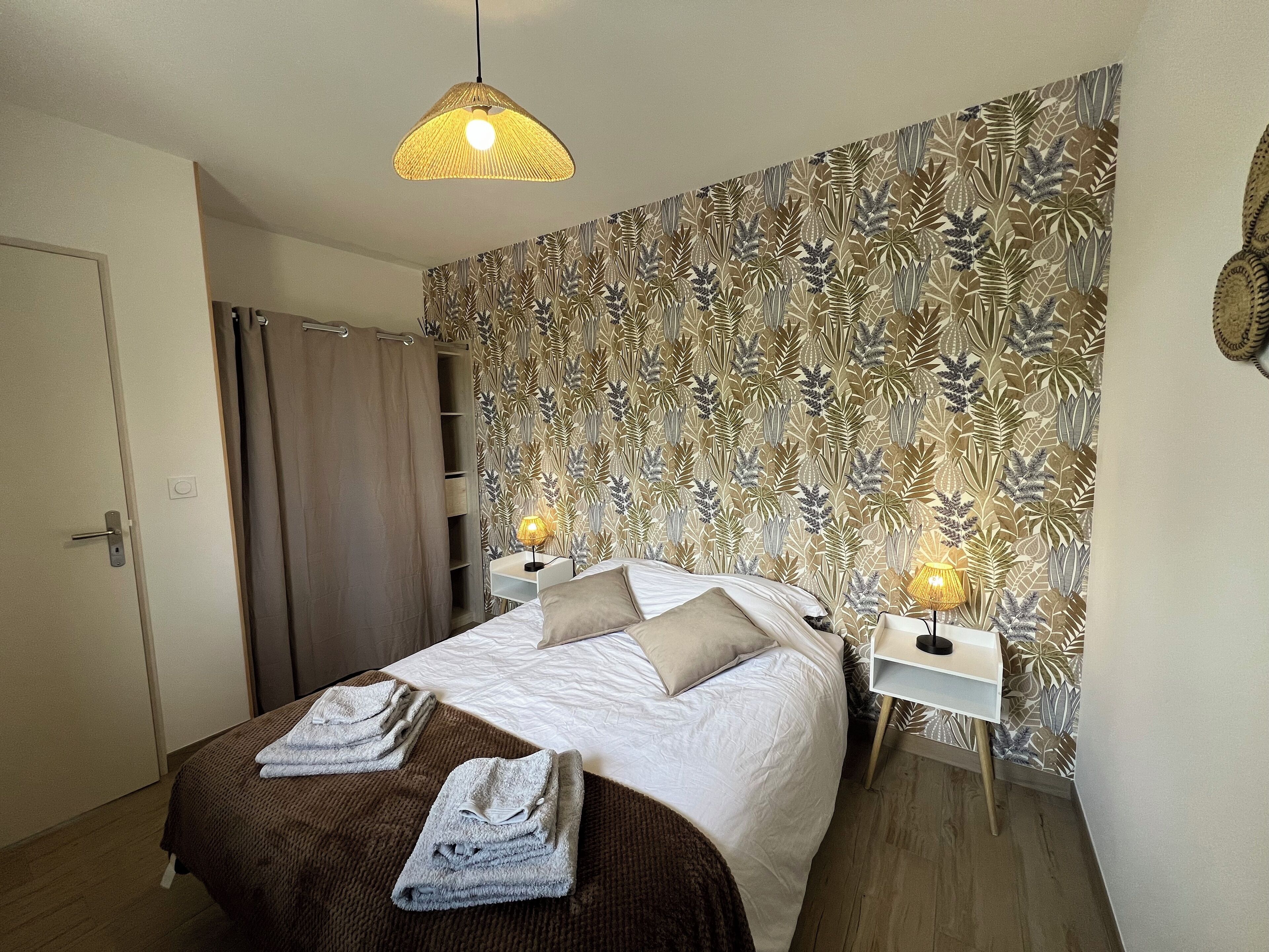 Een net opgemaakt bed met twee kussens, een nachtkastje met een lamp, en een muur met behang met bladmotieven.