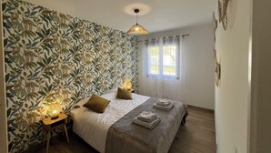 A bedroom with a patterned wallpaper, a bed with pillows, a bedside table with a lamp, and a window with curtains. - le Cocon Vézerontin (Vézeronce-Curtin)