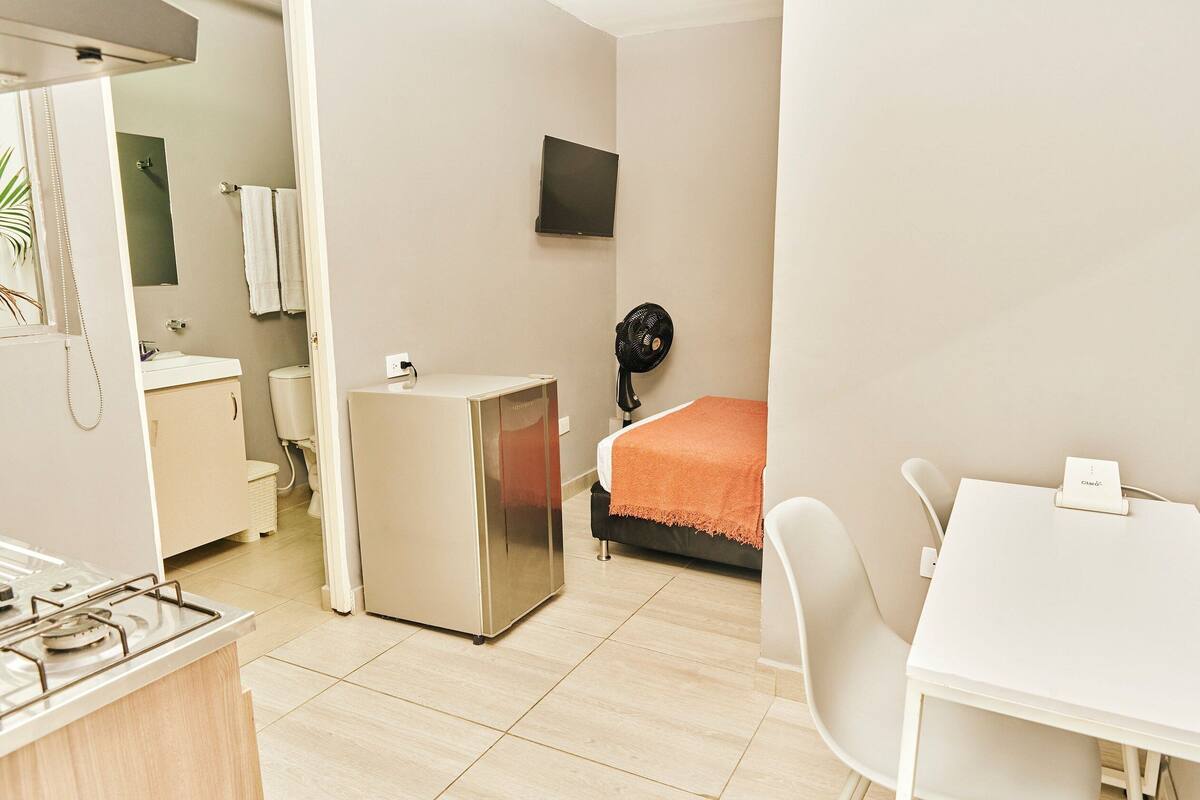 Ein kompaktes Hotelzimmer mit Küchenzeile, einem Bett mit orangefarbener Decke, einem an der Wand befestigten Fernseher und einem kleinen Ventilator.