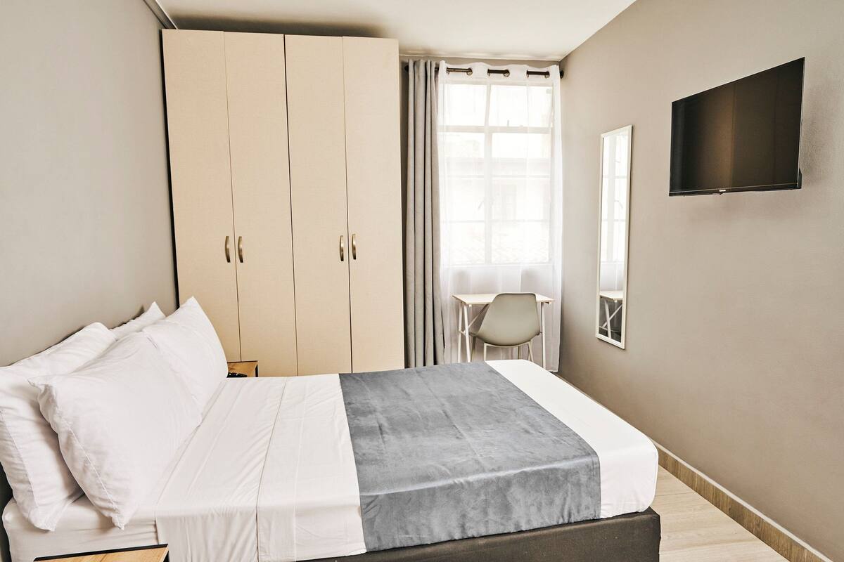 Ein Hotelzimmer mit Bett, Kleiderschrank, Schreibtisch, Stuhl und einem an der Wand befestigten Fernseher.