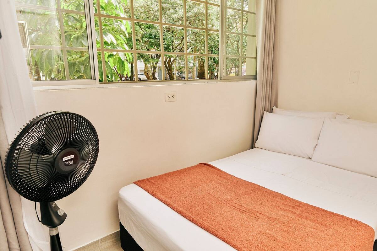 Ein Hotelzimmer mit Bett, Standventilator und einem Fenster mit Blick auf Grünflächen.