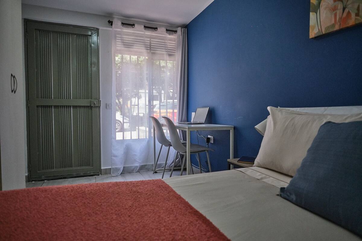 Ein Schlafzimmer mit einem Bett, einem Schreibtisch und einem Sessel. Ein Fenster mit Vorhängen bietet einen Blick nach draußen.