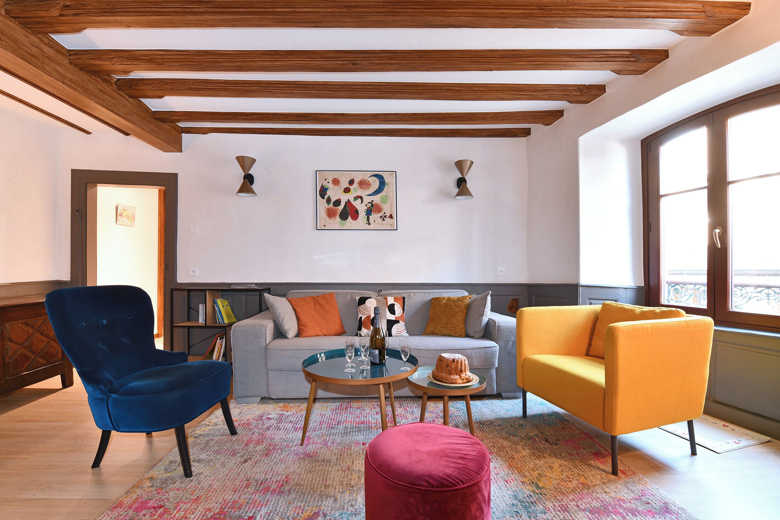 Un salon avec un fauteuil bleu, un canapé jaune, une table basse ronde et un pouf rouge. Il y a un tableau au mur et une fenêtre donnant sur un paysage.