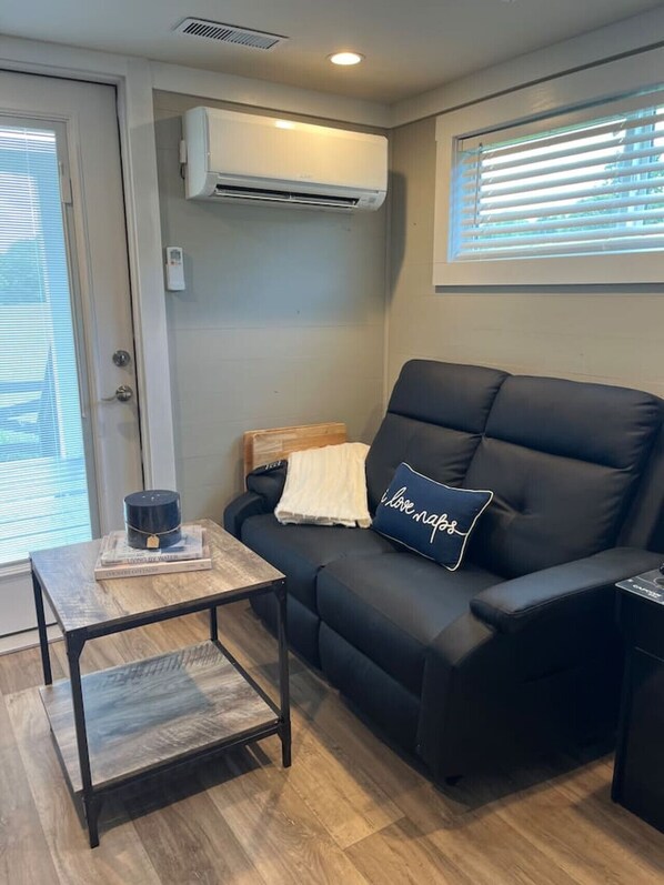 A modern living room with a black leather sofa, a small wooden coffee table, and a wall-mounted air conditioner. - Riverfront Tiny Home - Amazing Views (Strawberry Plains)