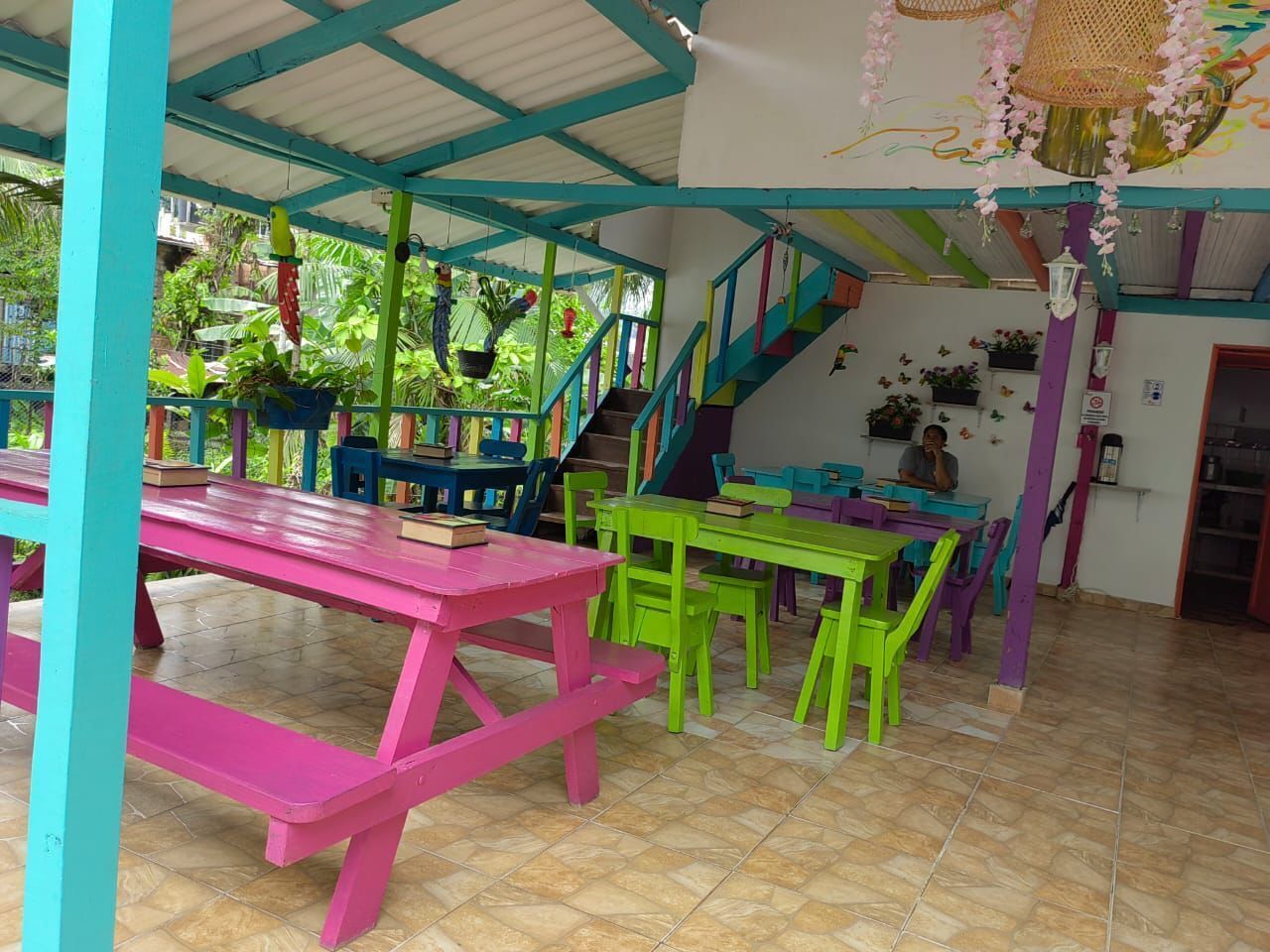 A brightly colored indoor space with multiple tables and chairs, a staircase, and a variety of plants.