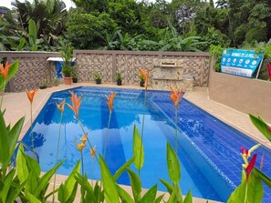 Une piscine dont le bord est recouvert de carrelage, entourée d’un mur de pierre et d’une végétation luxuriante.