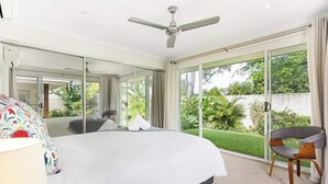 A modern bedroom with a large bed, a ceiling fan, and a view of a lush garden through large sliding doors. - Ocean55 Mooloolaba Holiday Home (Mooloolaba)