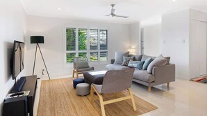 A modern living room with a sofa, chairs, a coffee table, a TV, and a floor lamp. - Ocean55 Mooloolaba Holiday Home (Mooloolaba)