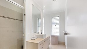 A modern bathroom with a glass shower enclosure, a large mirror, a vanity with a sink, and a window with a grid pattern. - Ocean55 Mooloolaba Holiday Home (Mooloolaba)
