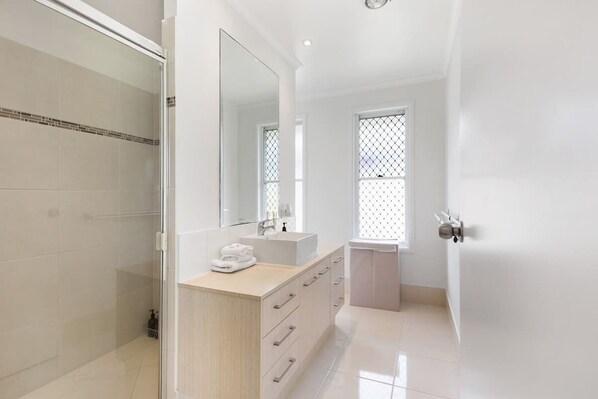 A modern bathroom with a glass shower enclosure, a large mirror, a vanity with a sink, and a window with a grid pattern. - Ocean55 Mooloolaba Holiday Home (Mooloolaba)