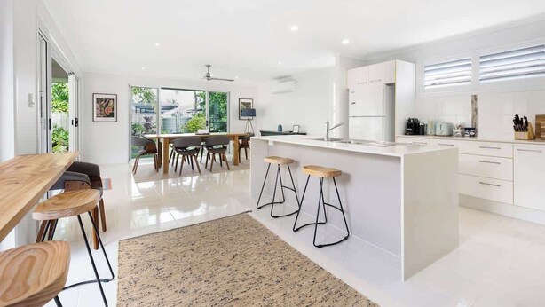A modern kitchen with white cabinetry, a central island with stools, and a dining area with a large table and chairs. - Ocean55 Mooloolaba Holiday Home (Mooloolaba)