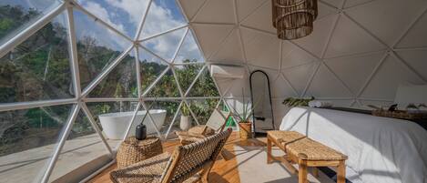 A spacious room with a large glass dome ceiling, wooden flooring, wicker furniture, a bed with white bedding, and a hanging light fixture.