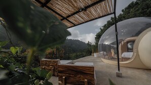 A transparent dome structure with a bed and seating area inside, surrounded by a wooden deck and plants. - Kafen Hotel  (Juayua)