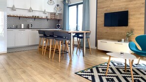A modern kitchen with a geometric backsplash, a dining area with a wooden table and chairs, a blue accent chair, and a mounted TV. - Modern Old Town 3 by Grand Apartments (Gdansk)