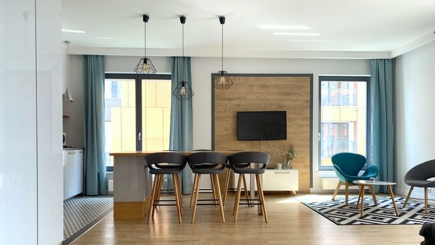A modern living room with a wooden floor, a dining area with four chairs, a TV mounted on the wall, and large windows with blue curtains. - Modern Old Town 3 by Grand Apartments (Gdansk)