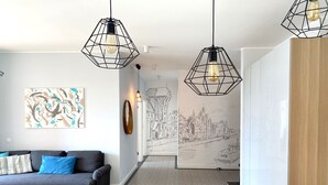 A modern living room with a grey sofa, black and blue cushions, a wooden coffee table, and two black chairs. There are three black pendant lights with geometric designs. A large abstract painting and a framed mirror are on the wall. A black and white cityscape mural is visible through an open door. - Modern Old Town 3 by Grand Apartments (Gdansk)