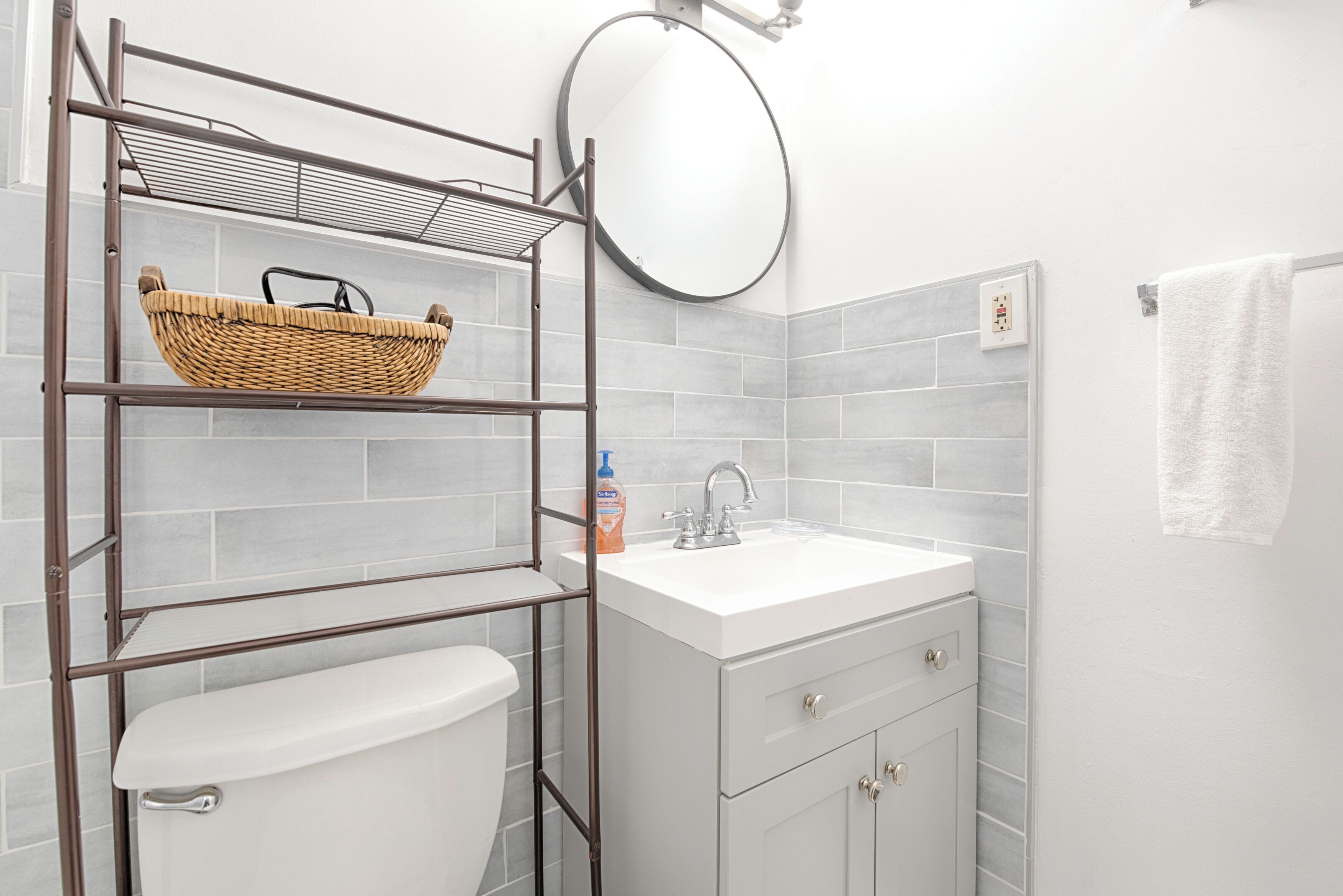 A bathroom with a white sink, a toilet, a wicker basket on a metal shelf, and a round mirror.