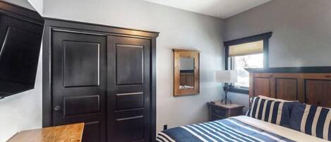 Un dormitorio con cama, una mesita de noche de madera, un armario oscuro, un espejo y una ventana con persianas.