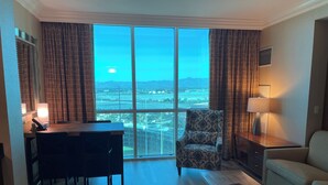 A spacious living room with a large window offering a city view, a comfortable sofa, a patterned armchair, a wooden coffee table, and a dining area with chairs and a lamp. - Renovated Luxury Studio Suite in Tower 3, New Luxury Suite Tower 3 (Paradise)