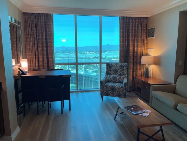 A spacious living room with a large window offering a city view, a comfortable sofa, a patterned armchair, a wooden coffee table, and a dining area with chairs and a lamp. - Renovated Luxury Studio Suite in Tower 3, New Luxury Suite Tower 3 (Paradise)
