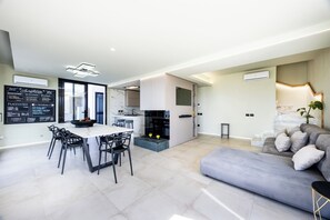 A modern living room with a grey sofa, a dining area with a table and chairs, and a kitchen area with a built-in stove.