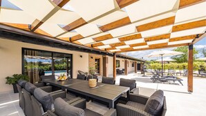 Une terrasse extérieure couverte, avec une table et des chaises, une piscine en arrière-plan et une vue sur le paysage environnant.