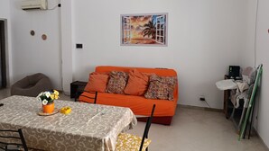 Een woonkamer met een bank, een eettafel met stoelen, een schilderij aan de muur en een bijzettafeltje met een bloevas.