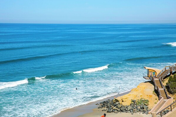A coastal beach with a wooden staircase leading to a yellow building, overlooking the ocean.