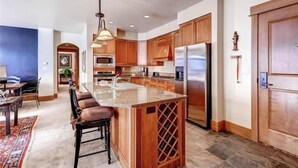 A modern kitchen with wooden cabinets, a central island, stainless steel appliances, and a dining area with a rug and chairs. - Ski in, ski out 3 Bedroom at Lone Eagle in River Run (Keystone)
