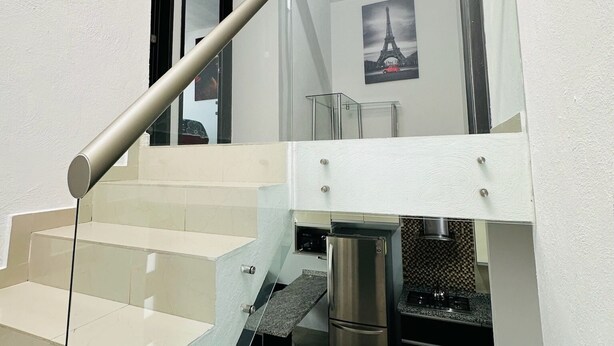 Una escalera moderna con barandillas de vidrio y un pasamanos metálico que conduce a una cocina con refrigerador de acero inoxidable y salpicadero azulejado.
