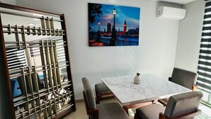 Un comedor con una mesa blanca y cuatro sillas grises tapizadas, un separador metálico decorativo y un cuadro de un paisaje urbano en la pared.