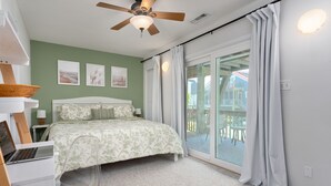A bedroom with a bed, a desk with a laptop, a ceiling fan, and a view of a balcony with chairs. - Paradise at The Point - Relaxing Home Steps to Beach + Sunsets + Ocean views (Holly Ridge)