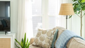 A living room with a beige sofa, a television on a white stand, a wicker floor lamp, a potted plant, and a window with sheer curtains. - Paradise at The Point - Relaxing Home Steps to Beach + Sunsets + Ocean views (Holly Ridge)