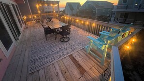 A wooden deck with a table and chairs, string lights, and a patterned rug. - Paradise at The Point - Relaxing Home Steps to Beach + Sunsets + Ocean views (Holly Ridge)