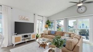 A modern living room with a large flat-screen TV, a beige sofa, a glass coffee table, and a white entertainment unit. - Paradise at The Point - Relaxing Home Steps to Beach + Sunsets + Ocean views (Holly Ridge)