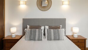 A neatly made bed with two pillows, a bedside table with a lamp, and a wall-mounted mirror with two wall sconces. - Serenidade do Mar & Spa (Peniche)