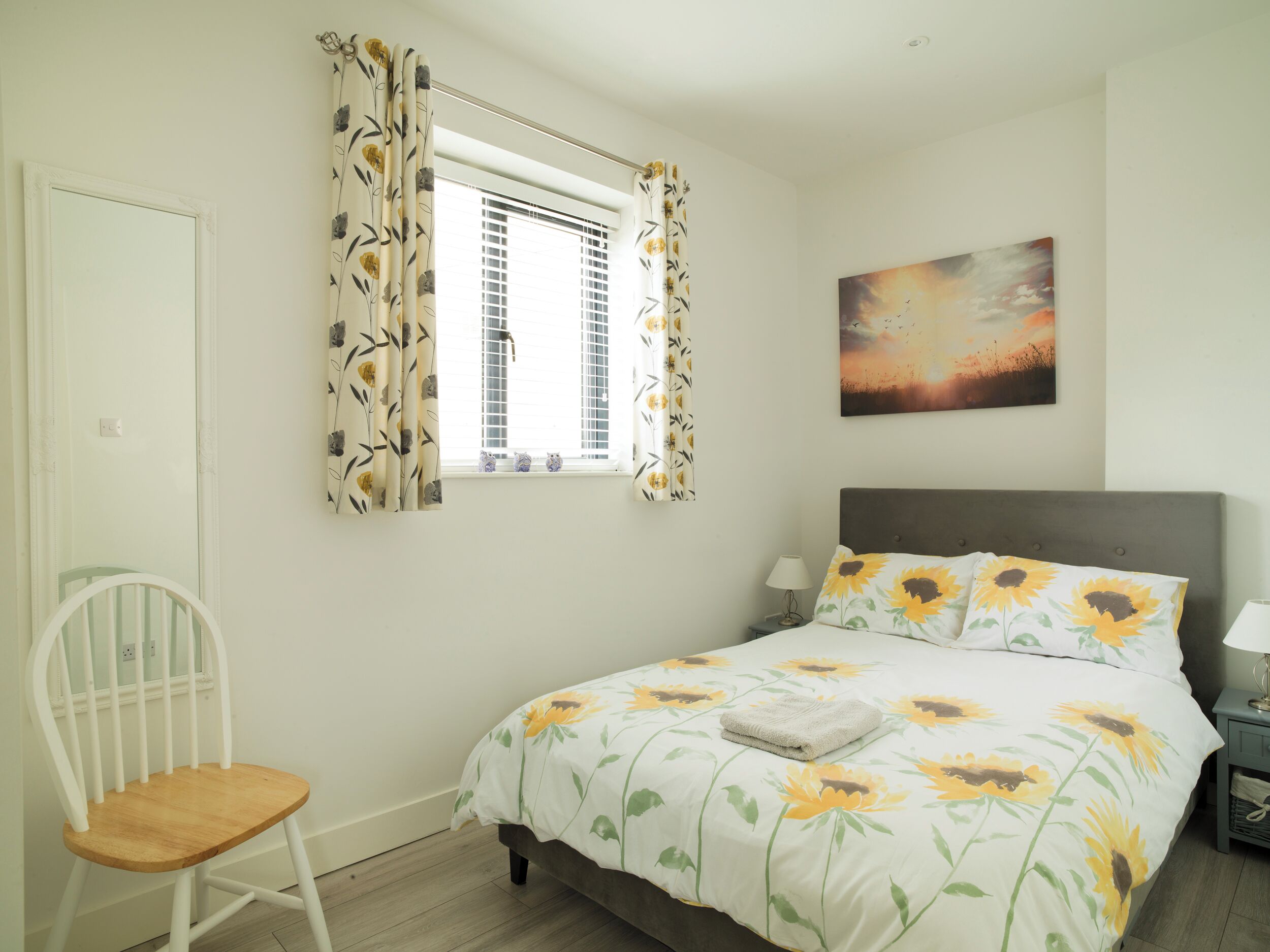 Une chambre à coucher avec un lit, une chaise, une fenêtre avec des rideaux, une table de chevet et un tableau au mur.