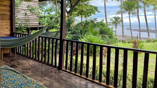 Een houten terras met een hangmat, een houten bankje en uitzicht op de oceaan en palmbomen.