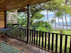 Una terraza de madera con una hamaca, un banco de madera y vistas al océano y palmeras.