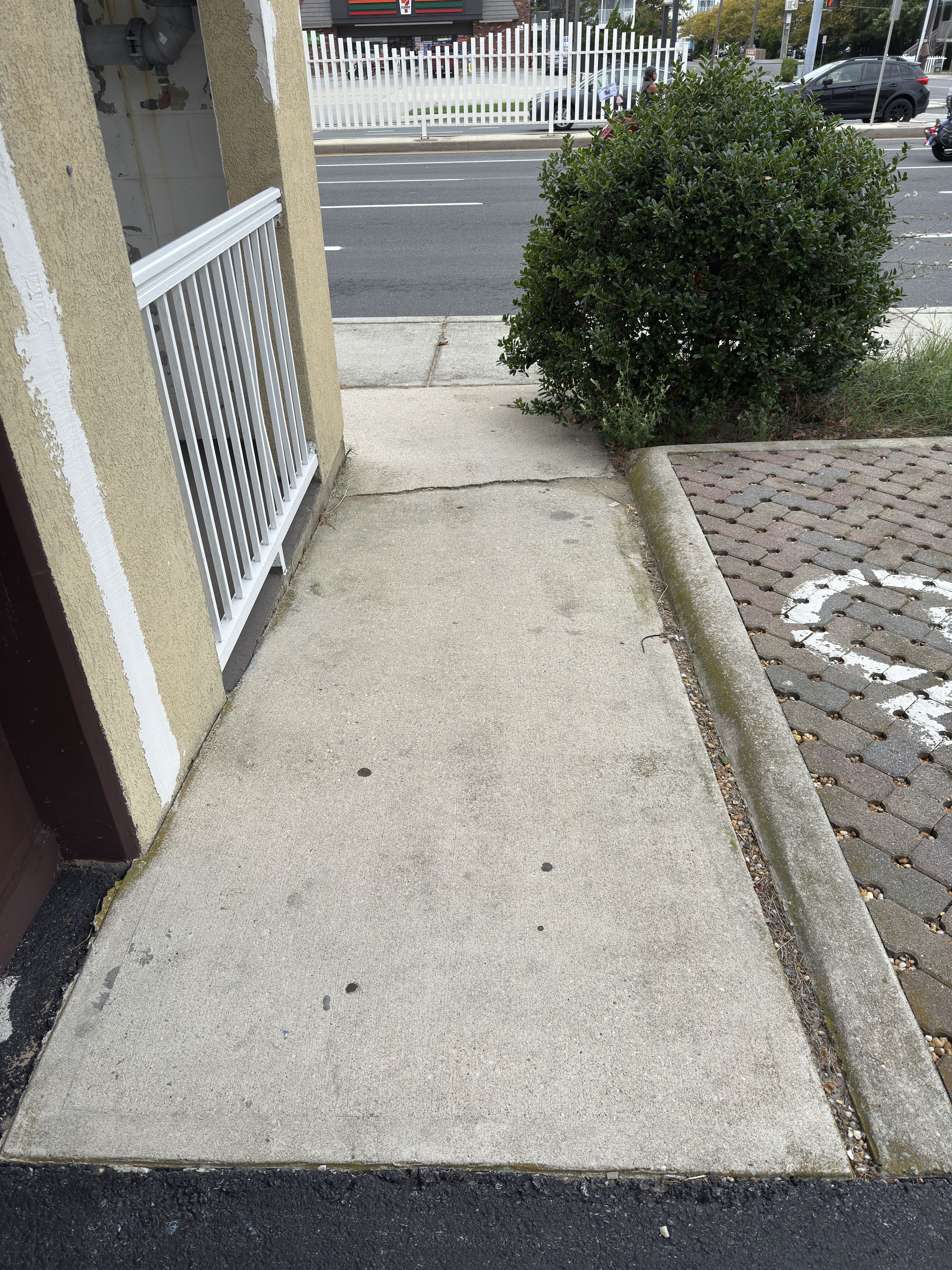 A concrete pathway leading to a building entrance with a white fence and a bush.