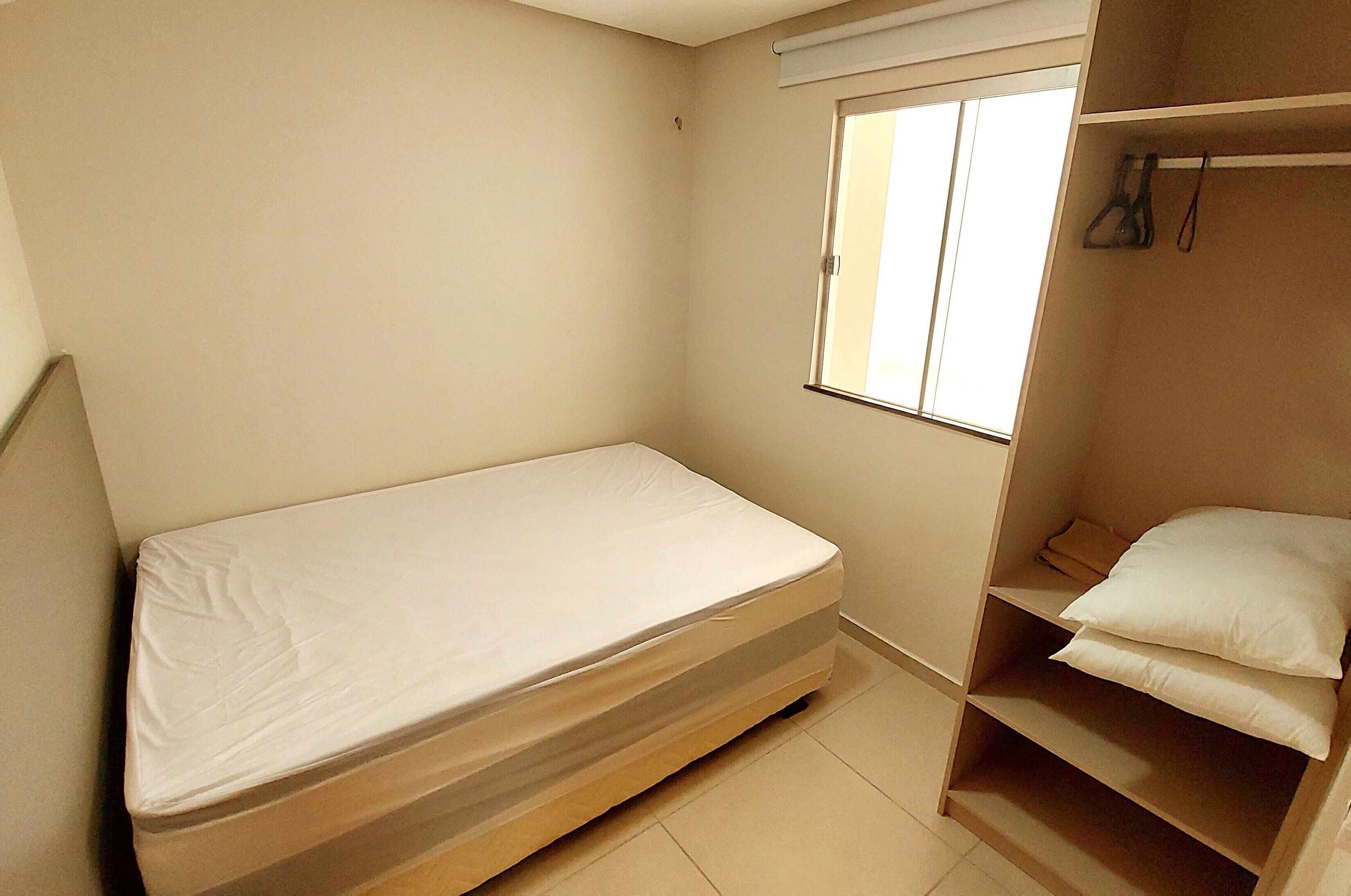 Una habitación pequeña con cama individual, ventana, armario y estante con almohadas.
